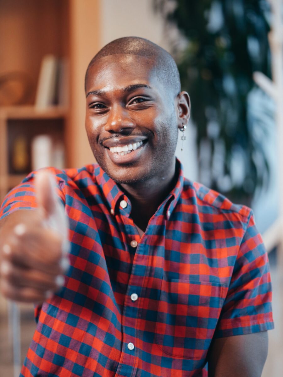 Portrait of a smiling man giving a thumbs up gesture in an indoor setting, wearing a checkered shirt.